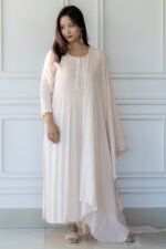 Full-length view of a woman wearing the blush pink Pavitra Anarkali kurti with pleated bodice, button accents, and matching dupatta, standing against a white paneled wall.
