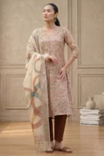 Person wearing a floral-patterned kurta with three-quarter sleeves, paired with brown churidar pants and a light dupatta with abstract multicolored designs, accessorized with hoop earrings, bracelet, and brown sandals, standing in a minimalist indoor setting.