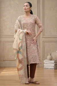 Person wearing a floral-patterned kurta with three-quarter sleeves, paired with brown churidar pants and a light dupatta with abstract multicolored designs, accessorized with hoop earrings, bracelet, and brown sandals, standing in a minimalist indoor setting.