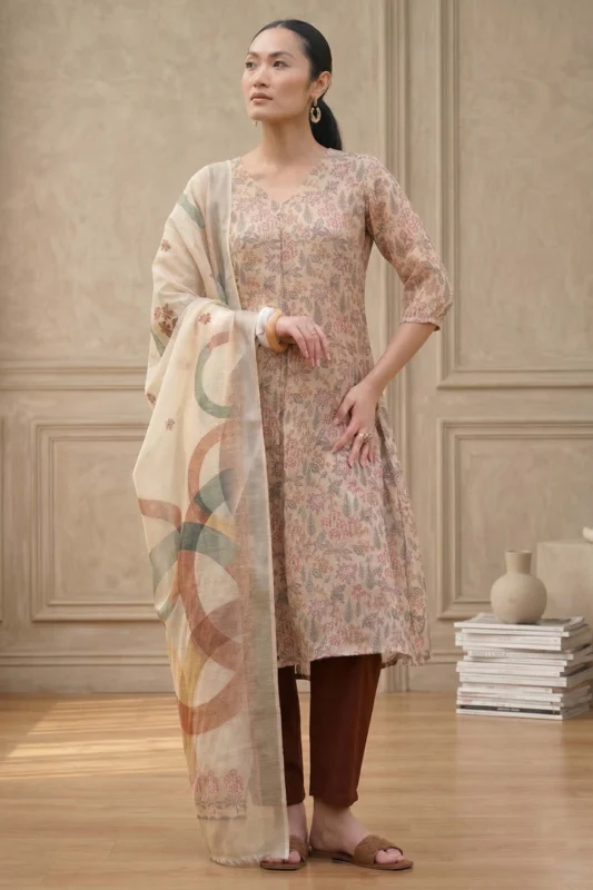 Person wearing a floral-patterned kurta with three-quarter sleeves, paired with brown churidar pants and a light dupatta with abstract multicolored designs, accessorized with hoop earrings, bracelet, and brown sandals, standing in a minimalist indoor setting.