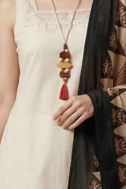 Close-up of a cream sleeveless dress with lace-trimmed square neckline, accessorized with a long necklace of multicolored wooden beads and red tassel, paired with a black and beige patterned shawl.