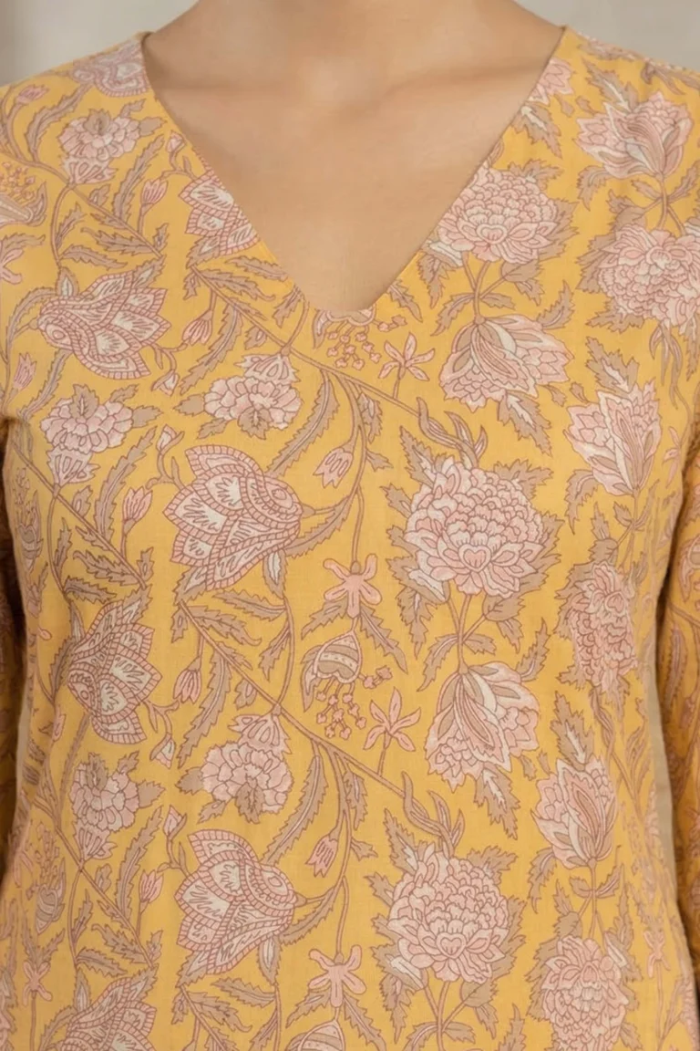 Close-up of a mustard yellow garment with a V-neckline, showcasing an intricate floral print in light pink and beige with repeating flowers and leaves.