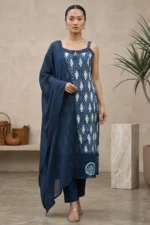 Person wearing a blue and white printed kurta with spaghetti straps, geometric and floral motifs, matching blue pants, and a blue dupatta, accessorized with earrings and beige heels, standing in a minimalist indoor setting with plant and woven baskets.