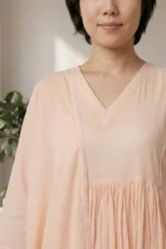 A detailed close-up of the ORA Ethnic V Neck peach kurti, showing the breathable fabric, gathered waistline, and delicate white lace trim on the sleeve cuffs.