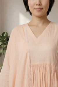 A detailed close-up of the ORA Ethnic V Neck peach kurti, showing the breathable fabric, gathered waistline, and delicate white lace trim on the sleeve cuffs.