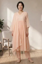 A full-length studio photo of a model in the ORA Ethnic V Neck peach three-piece set, featuring a flowy gathered kurta, straight trousers, and a lace-bordered dupatta.
