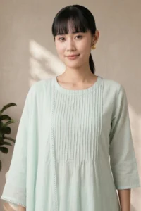 A portrait of a woman wearing the Nysa Weaves seafoam green kurti, highlighting the round neckline and the intricate vertical pin-tuck pleats on the bodice with a matching lace-trimmed dupatta.