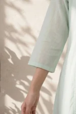 A detailed close-up of the Nysa Weaves kurti sleeve made from seafoam green cotton-linen fabric, showing the fine weave and clean hemline against a soft-focus background with leafy shadows.