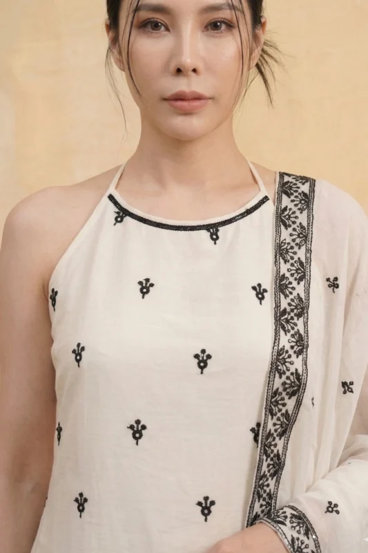 A close-up view of the StyleMélange kurta's bodice. The natural-toned fabric features a clean halter neckline accented with a thin black embroidered trim. Scattered black floral motifs decorate the chest, while a sheer dupatta with a dense black floral border is draped over the shoulder.