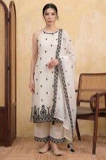 A woman with long dark hair stands in a studio wearing the StyleMélange monochrome suit set. The kurta is a natural kora beige with a contemporary halter-neck design and all-over black floral embroidery. It is styled with wide-leg beige palazzos and a sheer dupatta, both featuring heavy black embroidered borders and decorative tassels.