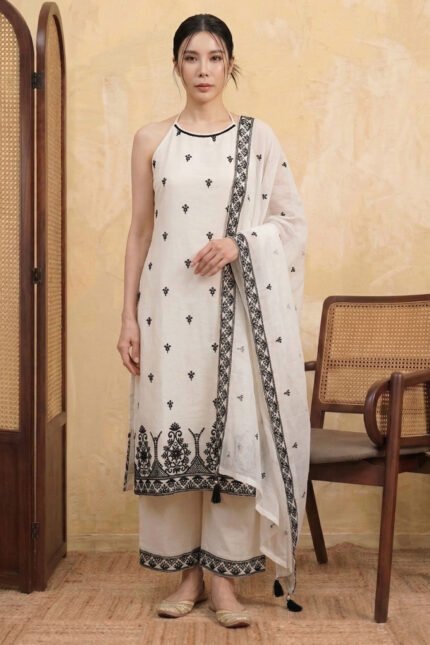 A woman with long dark hair stands in a studio wearing the StyleMélange monochrome suit set. The kurta is a natural kora beige with a contemporary halter-neck design and all-over black floral embroidery. It is styled with wide-leg beige palazzos and a sheer dupatta, both featuring heavy black embroidered borders and decorative tassels.