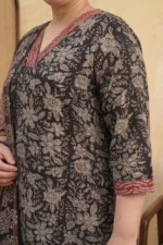 A woman stands smiling in a three-piece ethnic outfit from Mehak Magic. She is wearing a charcoal-colored, calf-length kurta with beige floral block prints and red patterned borders. The look is paired with black straight-fit trousers and a matching lightweight dupatta featuring chevron and paisley motifs.