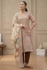 Person wearing a floral-patterned kurta with three-quarter sleeves, paired with brown churidar pants and a light dupatta with abstract multicolored designs, accessorized with hoop earrings, bracelet, and brown sandals, standing in a minimalist indoor setting.