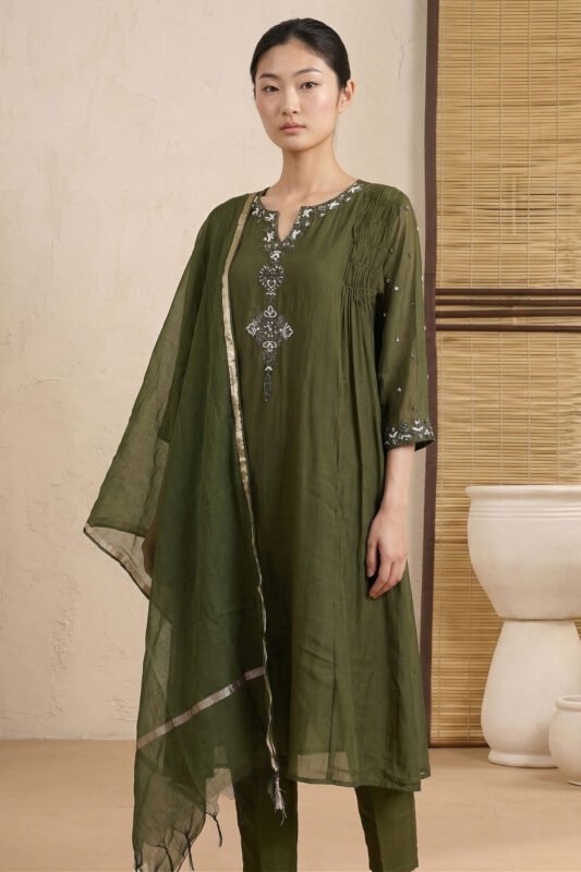 A full-length view of a woman wearing a vibrant crimson red kurti featuring vertical pintucks and white floral embroidery. She is standing in a minimalist room with soft natural lighting, paired with matching red trousers and a sheer white dupatta.