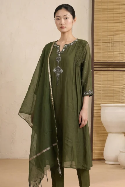 A full-length view of a woman wearing a vibrant crimson red kurti featuring vertical pintucks and white floral embroidery. She is standing in a minimalist room with soft natural lighting, paired with matching red trousers and a sheer white dupatta.