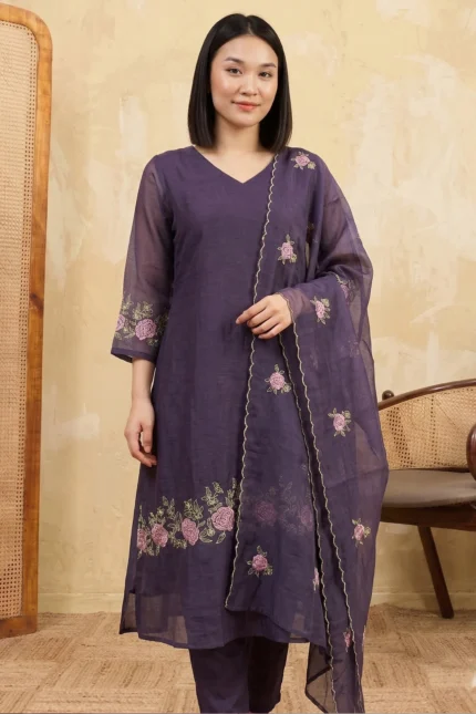 A full-length view of a woman wearing an elegantly tailored long kurti from the Lotus Line collection. The garment features floral embroidery along the hem and is paired with matching straight-cut trousers and a sheer dupatta in a minimalist indoor setting.