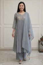 A full-length view of a woman wearing a teal-colored flared kurti from the Majuli Muse collection. The garment features intricate floral embroidery on the bodice and hem, paired with matching teal trousers and a sheer dupatta.