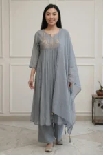A full-length view of a woman wearing a teal-colored flared kurti from the Majuli Muse collection. The garment features intricate floral embroidery on the bodice and hem, paired with matching teal trousers and a sheer dupatta.