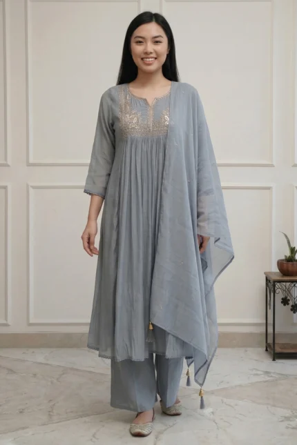 A full-length view of a woman wearing a teal-colored flared kurti from the Majuli Muse collection. The garment features intricate floral embroidery on the bodice and hem, paired with matching teal trousers and a sheer dupatta.