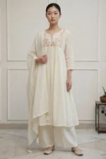 A full-length view of a woman wearing an all-white flared kurti from the Majuli Muse collection. The garment features intricate white-on-white floral embroidery on the bodice and a wide embroidered border at the hem, paired with matching white trousers and a sheer dupatta.