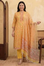A woman stands smiling in a bright sun-yellow Mehak suit set. The knee-length kurta is made of airy Kota Doria fabric featuring an all-over orange and pink floral block print. She wears solid yellow straight-cut trousers and a matching sheer floral dupatta with tassel details. The outfit is paired with gold ethnic sandals.