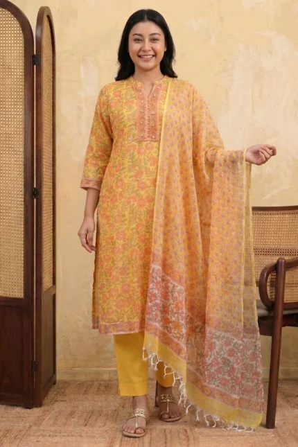 A woman stands smiling in a bright sun-yellow Mehak suit set. The knee-length kurta is made of airy Kota Doria fabric featuring an all-over orange and pink floral block print. She wears solid yellow straight-cut trousers and a matching sheer floral dupatta with tassel details. The outfit is paired with gold ethnic sandals.