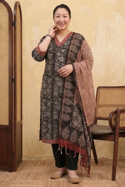 Full-length view of a Mehak Magic floral print kurta set. The garment features three-quarter sleeves, a V-neckline with red piping, and a coordinated patterned dupatta, styled with a cane room divider and wooden chair in the background.