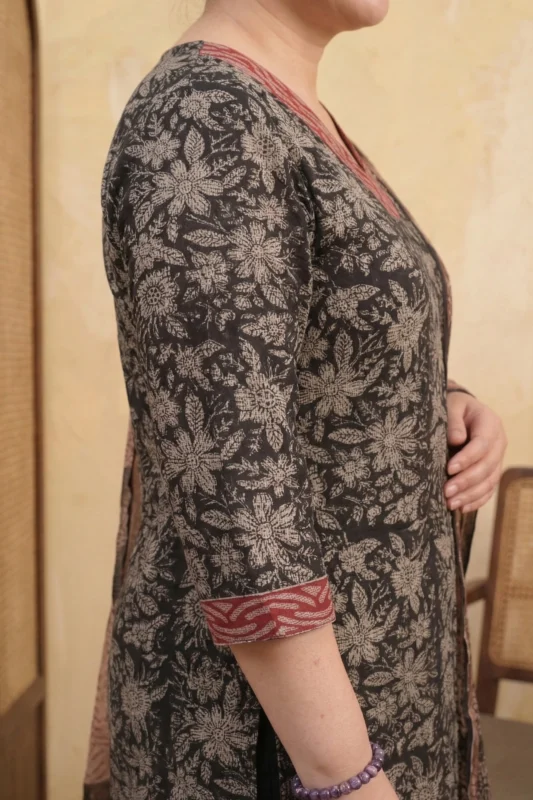 A close-up and full shot of a Mehak Magic ethnic ensemble. The charcoal fabric is detailed with intricate beige floral patterns. The model is accessorized with a purple beaded bracelet and traditional jutti shoes, posing in a warm, neutral-toned room.