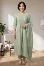 A full-length view of the Nazm sage green kurta set. The model is wearing a long-sleeved A-line kurta with pleated details, matching straight-fit trousers, and a soft organza dupatta with white lace trim.