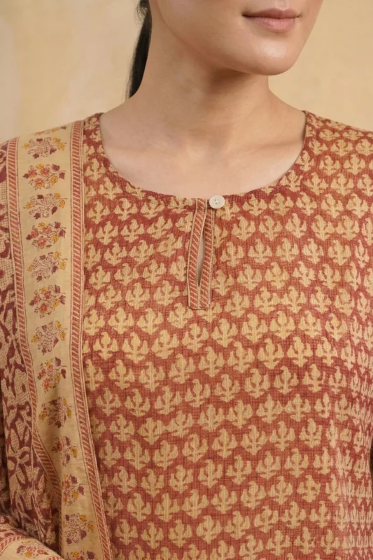 A close-up view of the Nysa kurta’s bodice. The design shows a simple round neckline with a small keyhole opening and a single white button. The rust-colored Kota Doria fabric displays a repeating hand-block motif in a lighter cream shade. A portion of the matching dupatta with a floral border is draped over the shoulder.
