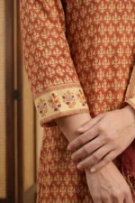 A detailed shot of the Nysa kurta sleeve against a warm background. The lightweight rust Kota Doria fabric features a consistent block print, finishing with a cream-toned border cuff. The cuff is decorated with hand-block printed floral sprigs in orange and purple tones, framed by thin striped borders.