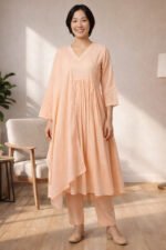 A full-length studio photo of a model in the ORA Ethnic V Neck peach three-piece set, featuring a flowy gathered kurta, straight trousers, and a lace-bordered dupatta.