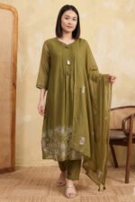 A woman stands wearing a flowy, olive green long kurti with white floral embroidery at the hem. She is paired with matching trousers and a sheer dupatta, styled against a minimalist beige background with a wooden room divider.