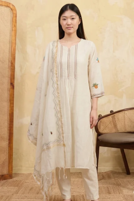 A full-length view of a woman wearing a sophisticated kurti set from the Regal Wear collection. The outfit features delicate floral embroidery along the lower hem and sleeve cuffs, paired with matching trousers and a lightweight dupatta in a bright, minimalist setting.