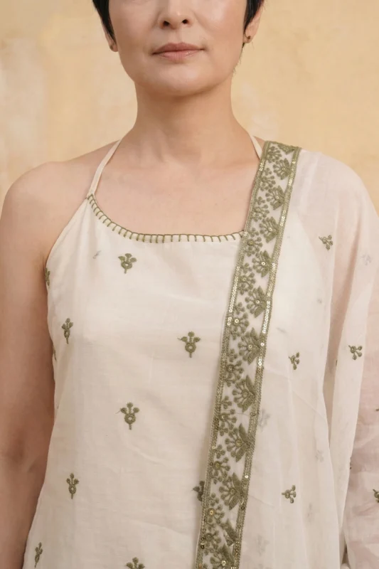 A close-up view of the StyleMélange kurta's upper bodice. The natural beige fabric is adorned with small, evenly spaced olive green floral motifs. The square neckline is finished with delicate green hand-embroidery. A sheer dupatta with a heavy olive green embroidered border and subtle sequin accents is draped over one shoulder, highlighting the earthy color palette.