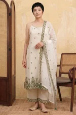 A woman with short dark hair stands in a neutral-toned studio wearing the StyleMélange suit set in a natural kora beige shade. The kurta features a unique halter-style neckline with thin straps and all-over olive green floral embroidery. It is paired with matching wide-leg beige palazzos and a sheer dupatta, both featuring intricate olive embroidered borders. She completes the look with traditional gold-toned juttis.