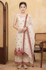 A woman stands in a studio wearing the StyleMélange maroon suit set. The kurta is a natural kora beige with a contemporary halter-neck design and scattered maroon floral embroidery. It is styled with matching wide-leg beige palazzos and a sheer dupatta, both featuring heavy maroon embroidered borders and decorative tassels at the corners.