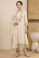 A full-length view of a woman wearing a flared Anarkali-style kurti with vibrant floral embroidery across the bodice, hem, and sleeve cuffs. The ensemble includes matching embroidered trousers and a coordinating dupatta, styled with traditional flats in a minimalist interior.