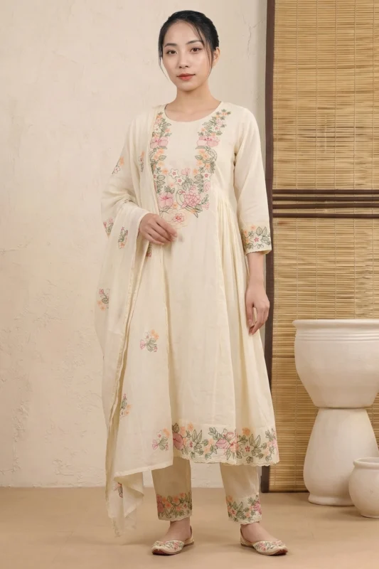 A full-length view of a woman wearing a flared Anarkali-style kurti with vibrant floral embroidery across the bodice, hem, and sleeve cuffs. The ensemble includes matching embroidered trousers and a coordinating dupatta, styled with traditional flats in a minimalist interior.