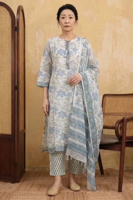 A woman stands in a warm-toned studio wearing the Urban Utopia suit set. The outfit is crafted from lightweight white Kota Doria fabric, featuring delicate blue and green floral block prints. The ensemble includes a knee-length kurta with tassel ties, straight-fit printed trousers, and a matching sheer dupatta draped over one shoulder.