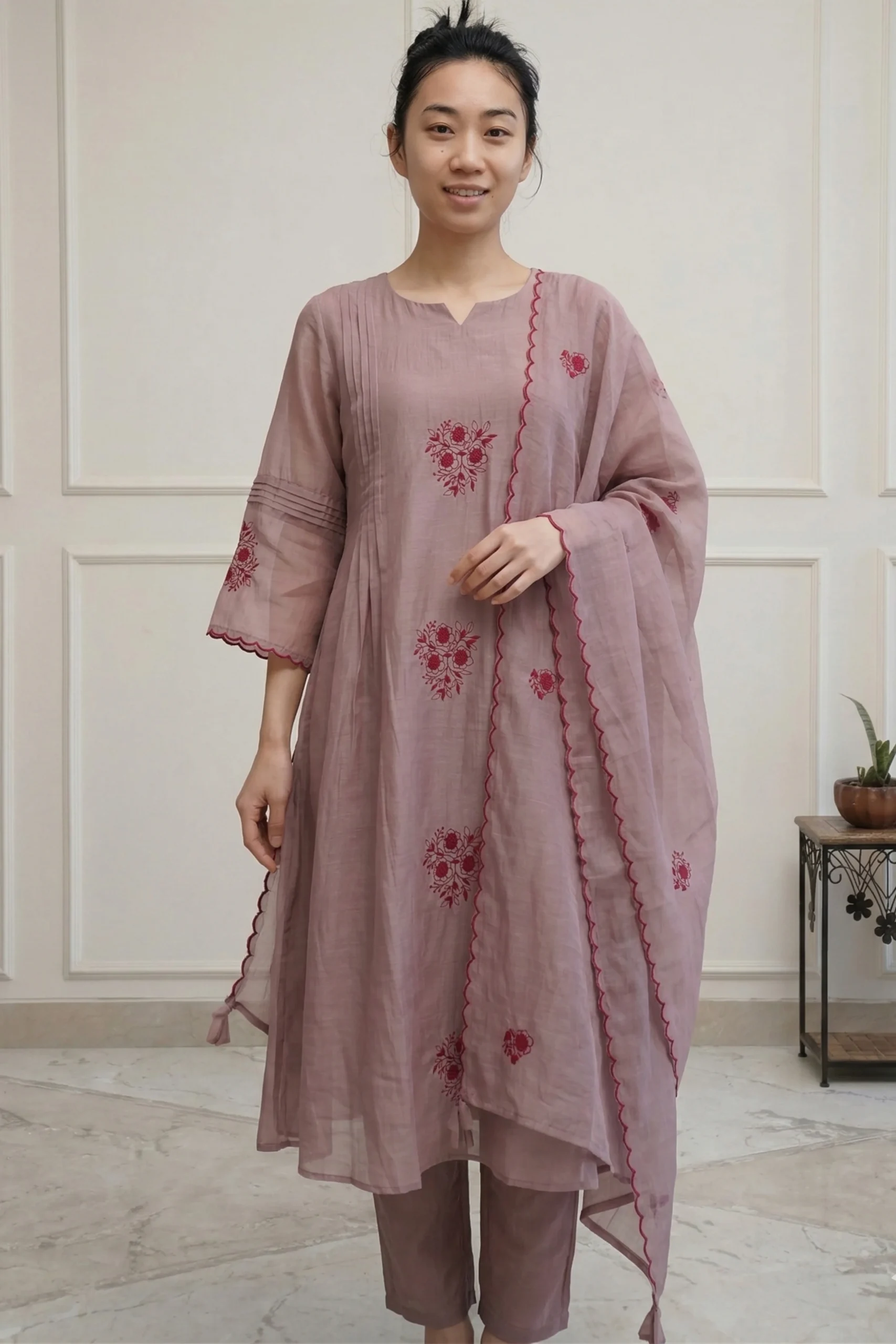 Velvet Veins Hand-Embroidered Designer Kurti Set A full-length view of a woman wearing a long, elegantly tailored kurti with intricate floral embroidery at the hem. She is styled with matching straight-fit trousers and a sheer dupatta, standing in a brightly lit, minimalist interior.