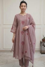 A full-length view of a woman wearing a long, elegantly tailored kurti with intricate floral embroidery at the hem. She is styled with matching straight-fit trousers and a sheer dupatta, standing in a brightly lit, minimalist interior.