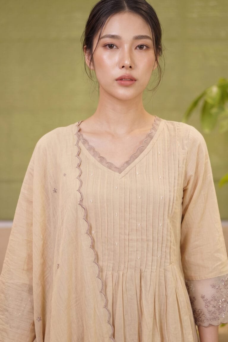 Close-up front view of The Dune Kurti in natural kora beige, highlighting the V-neckline with scalloped lace trim and delicate pintuck pleating across the chest.