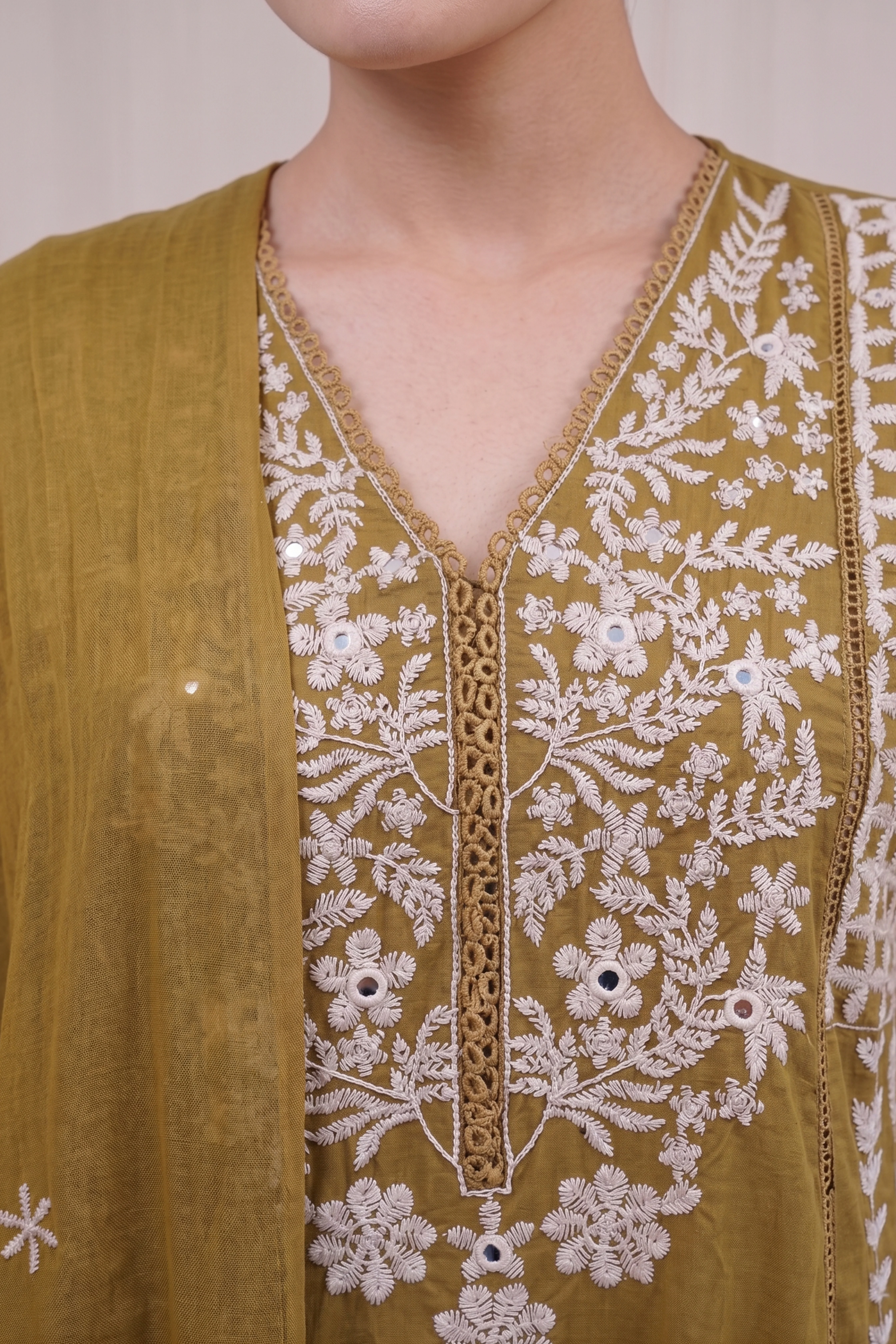 Close-up of the V-neckline of the Zoya kurti showing intricate white thread work, floral embroidery motifs, and delicate mirror work accents on a mustard olive fabric.