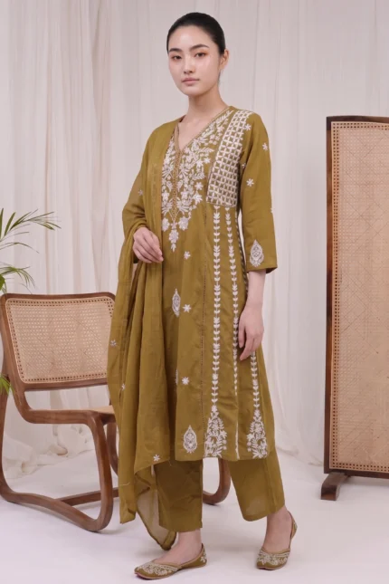 A full-length shot of a model wearing the Zoya olive green ethnic suit set, featuring white floral embroidery, matching straight trousers, and a dupatta, styled with embroidered juttis in a minimalist studio setting.