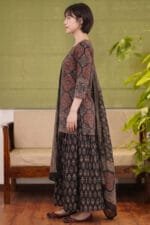 Full-length side profile view of a model wearing the dark-based Midnight Halo Kurti set, highlighting the straight-cut hem and intricate red circular motifs. The look is paired with a matching flared bottom showcasing repeating botanical prints and a draped patterned dupatta, set against a minimal studio background featuring a cane-weaved wooden sofa and an indoor plant.