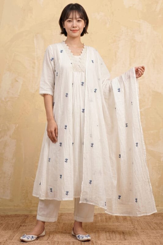 Full-length, front-angled view of a model wearing the crisp white Pearl Gather Kurti set. The A-line silhouette features scattered Dusty Blue embroidered floral motifs, a prominent lace-detailed yoke, and coordinated straight pants with delicate scalloped lace hems. The look is finished with a matching draped dupatta against a minimal studio background featuring a textured beige wall and a woven floor mat.