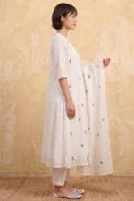 Full-length side profile view of a model wearing the Pearl Gather Kurti set, highlighting the soft, gathered A-line silhouette and the precise alignment of the lace detailing on the sleeve hem. The crisp white ensemble, dotted with Dusty Blue motifs, is paired with matching straight pants and a draped dupatta, captured within a premium minimal studio setting.