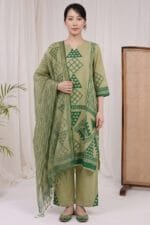 Full-length front view of a model wearing the Sage Geo Kurti set, featuring a straight-cut silhouette and matching straight trousers with prominent triangular border prints at the hem. The draped dupatta displays repeating geometric and botanical motifs, set against a minimal studio background with soft beige curtains, a cane-weaved wooden divider, and a subtle indoor plant.