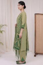 Full-length side profile view of a model in the Sage Geo Kurti set, highlighting the straight hemline, precise side slit alignment, and coordinated geometric print on the trousers. The draped dupatta falls elegantly at the side, captured within a premium studio aesthetic featuring beige curtains and minimal wooden cane elements.